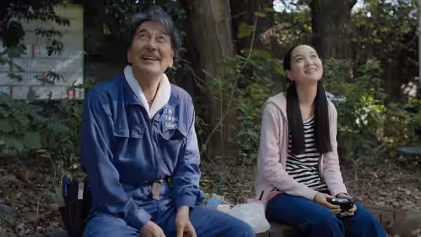 Hirayama (Koji Yakusho) and a young woman sit on a bench in the woods looking up