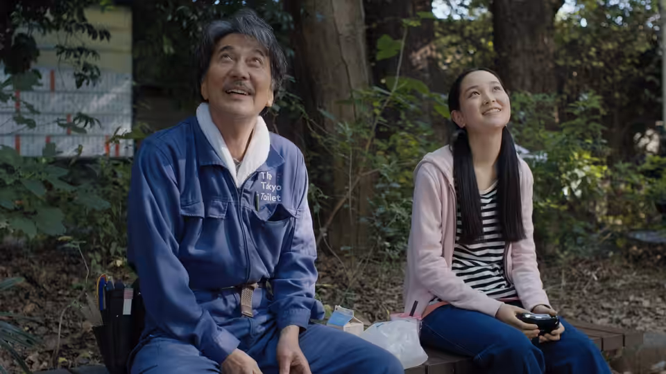 Hirayama (Koji Yakusho) and a young woman sit on a bench in the woods looking up