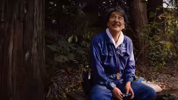 Hirayama (Koji Yakusho) sits on a bench in the forest holding a video game controller