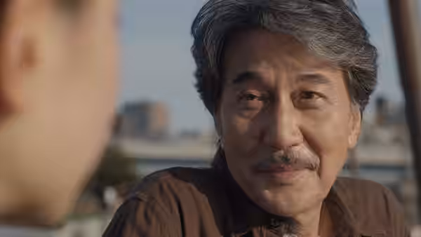 Hirayama (Koji Yakusho) smiles slightly while looking at someone outdoors with a city view behind
