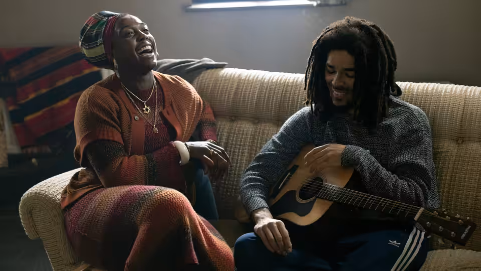 Rita Marley (Lashana Lynch) laughing beside Bob Marley (Kingsley Ben-Adir) holding guitar on couch