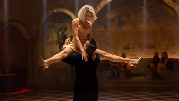 A blonde woman in a gold dress is lifted horizontally by a man in a dark shirt