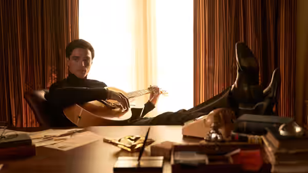 Elvis (Jacob Elordi) playing guitar with feet propped on a desk