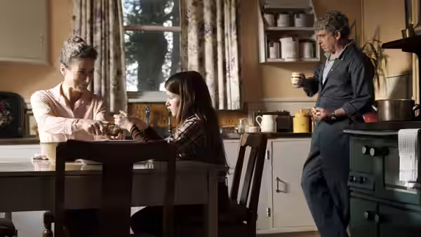 Cáit (Catherine Clinch) sits at a kitchen table while Eibhlín (Carrie Crowley) pours a drink and Seán (Andrew Bennett) watches