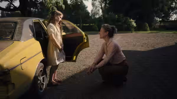 Cáit (Catherine Clinch) stands beside a yellow car as Eibhlín (Carrie Crowley) crouches to speak with her