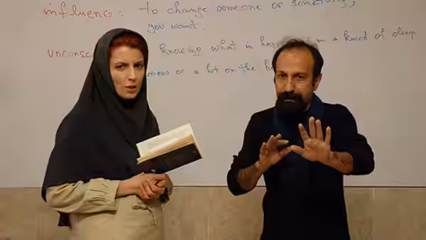 A woman holding an open book stands next to a man gesturing before a whiteboard