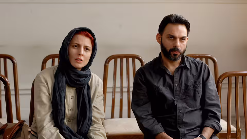 A woman in a headscarf sits beside a bearded man on wooden chairs in a hall
