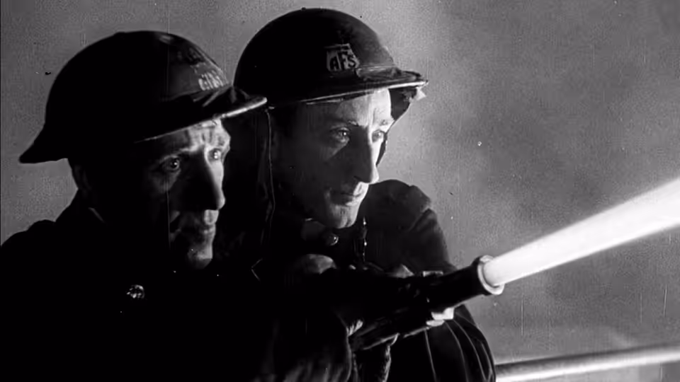Two firemen in helmets brace a hose nozzle inside a darkened building