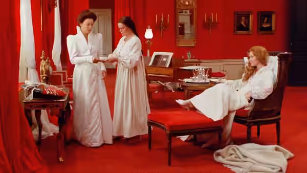 two women in white stand facing each other near a third reclining in a chair