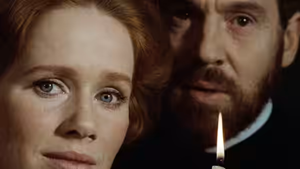 a red-haired woman and a bearded man stand close beside a lit candle