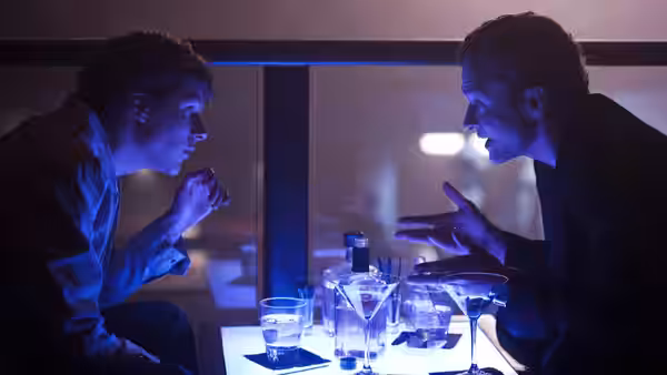 Jesse Eisenberg faces Justin Timberlake across a table with cocktails in blue light