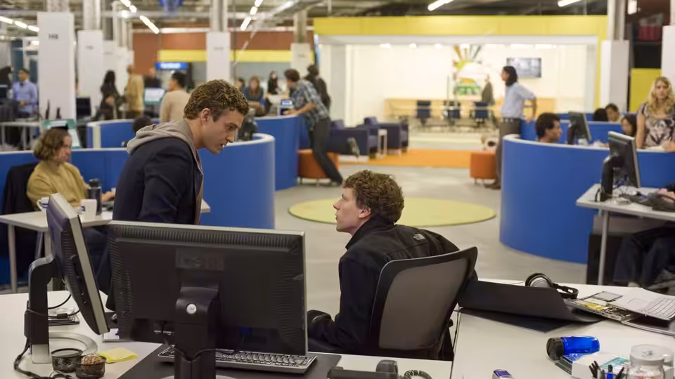 Andrew Garfield stands speaking to a seated colleague in a busy open office