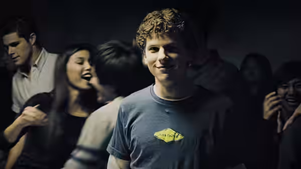 A smiling young man in a blue t-shirt stands at a crowded indoor party