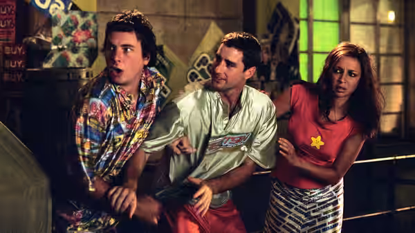 Joe Bauers (Luke Wilson) gesturing as Frito (Dax Shepard) and Rita (Maya Rudolph) react with surprise indoors