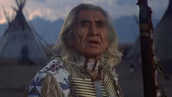 Old Lodge Skins (Chief Dan George) stands in traditional dress beside a tent