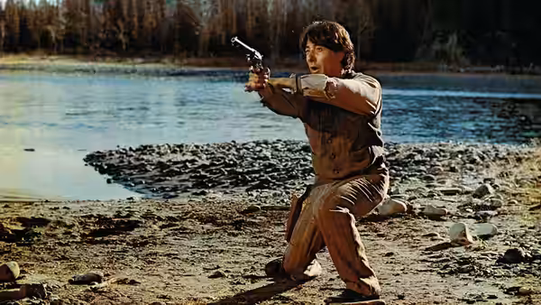 Jack Crabb (Dustin Hoffman) kneels on a lakeshore, aiming a revolver