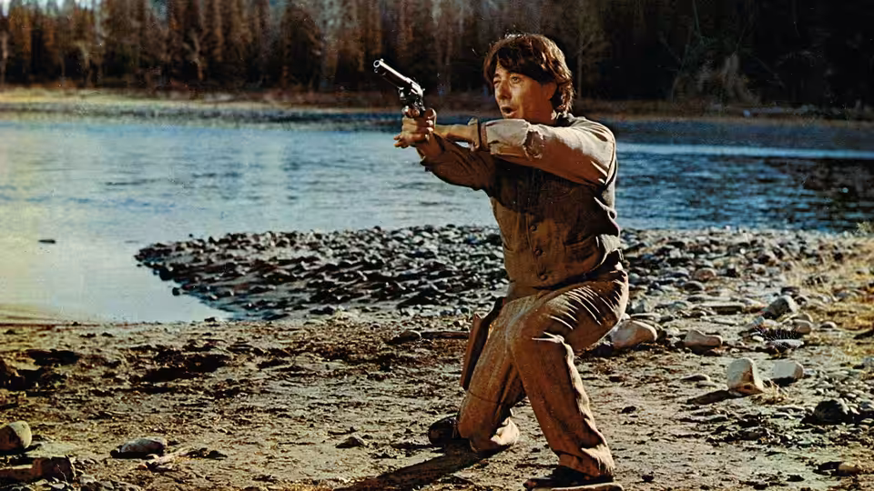 Jack Crabb (Dustin Hoffman) kneels on a lakeshore, aiming a revolver
