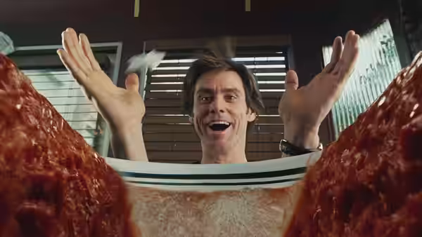 Bruce Nolan (Jim Carrey) raising both hands behind a massive sandwich at a desk