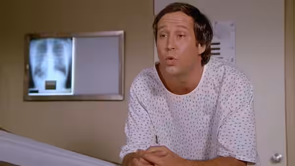 Fletch leans over a hospital bed in a patient gown