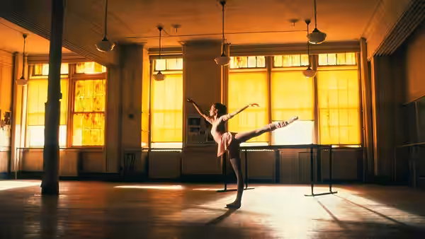A ballet dancer balances with one leg raised in a sunlit studio
