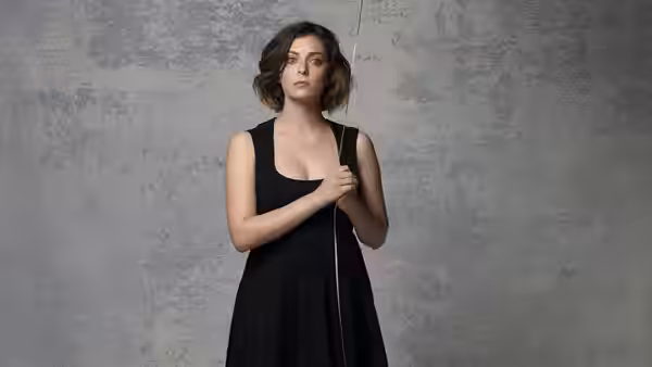Rachel Bloom in a black dress holding a cord against a gray wall