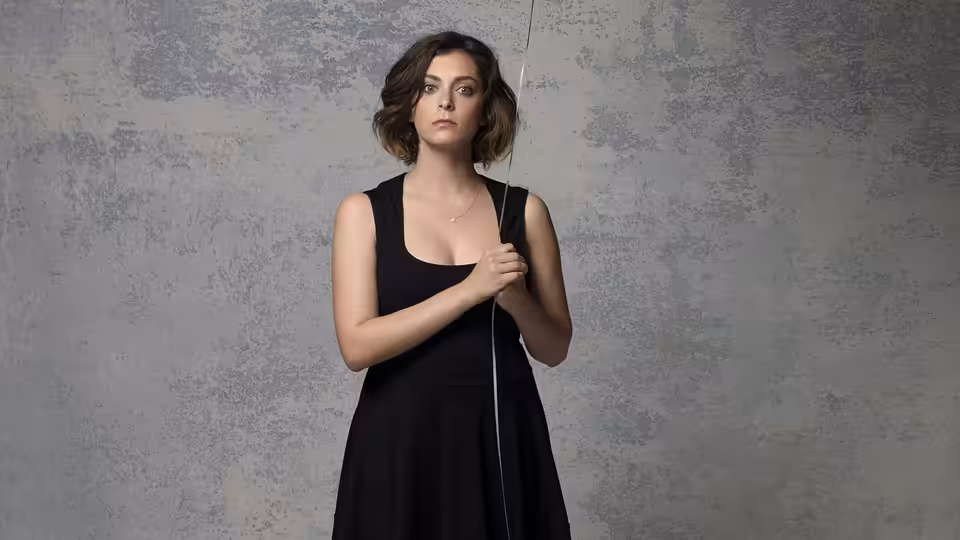 Rachel Bloom in a black dress holding a cord against a gray wall