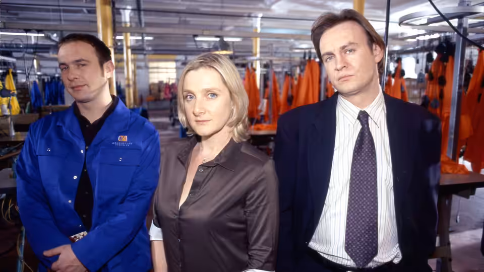 Three adults stand among garment racks on a factory floor