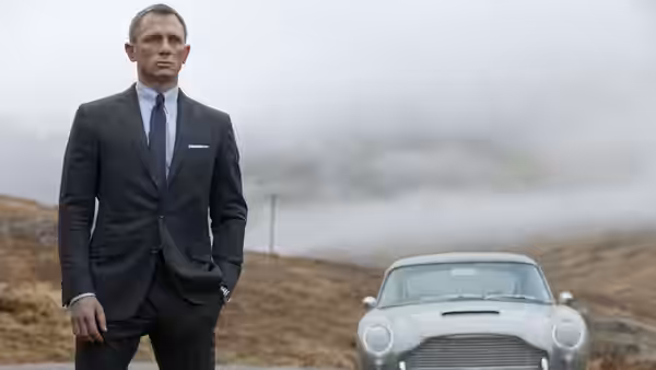 James Bond (Daniel Craig) standing with hands in pockets beside a silver sports car