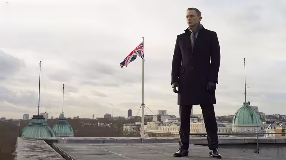 James Bond (Daniel Craig) in a dark overcoat on a rooftop with a Union Jack flag