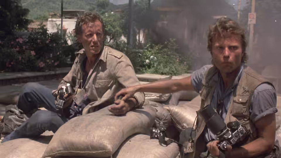 Richard Boyle (James Woods) and Doctor Rock (Jim Belushi) crouch behind sandbags with cameras