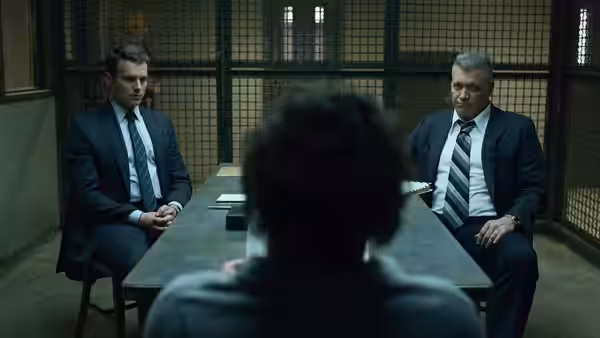 Two suited men seated at a table facing a person in an interview room
