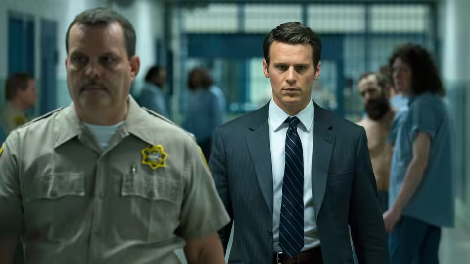 Holden Ford beside a sheriff's deputy walking through a prison hallway