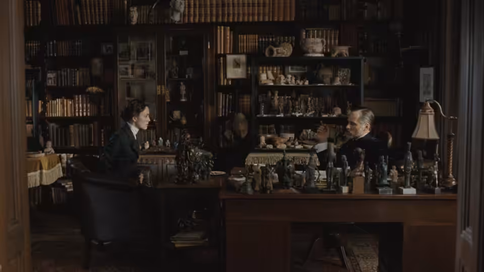 A woman and a man sit across from each other in a book-lined study
