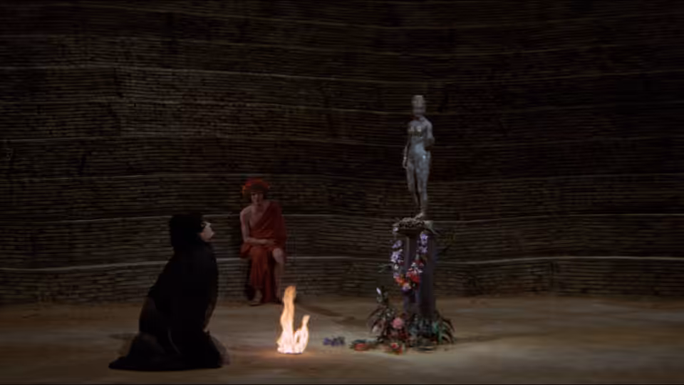 A black-robed figure watches a small fire as another person stands atop a decorated structure