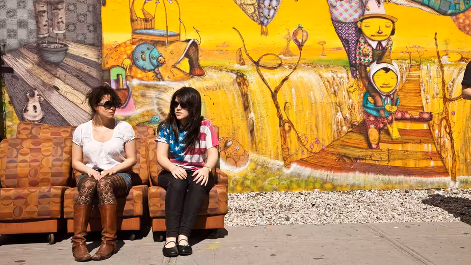Abbi Abrams (Abbi Jacobson) and Ilana Wexler (Ilana Glazer) seated on an orange couch before a colorful mural