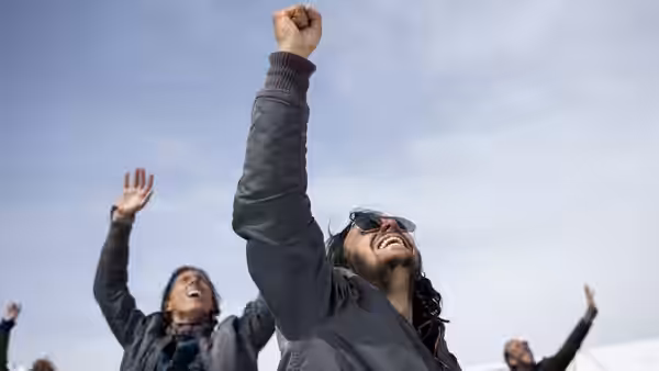 A bearded man and fellow survivors raise their arms beneath a cloudy sky