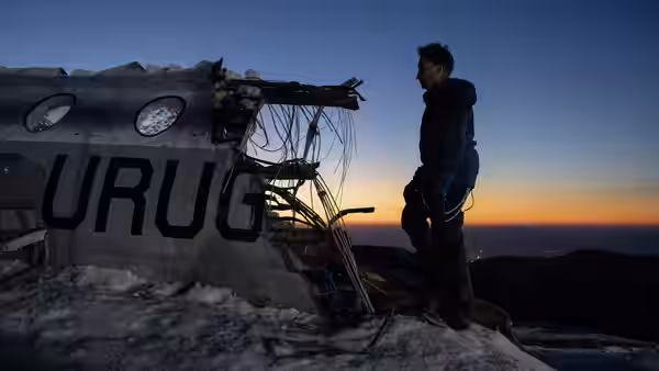 A man stands beside the wreckage of a plane on a snowy ridge at sunset