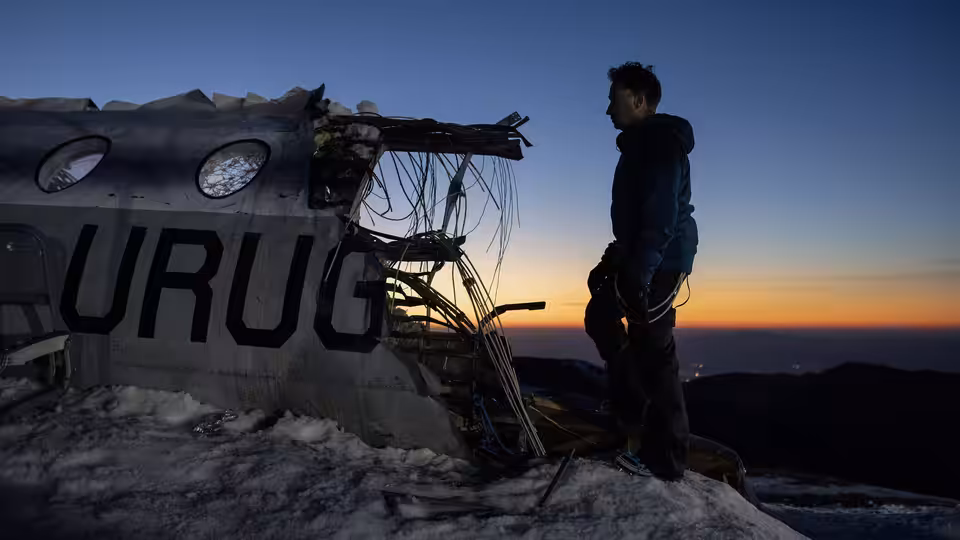 A man stands beside the wreckage of a plane on a snowy ridge at sunset