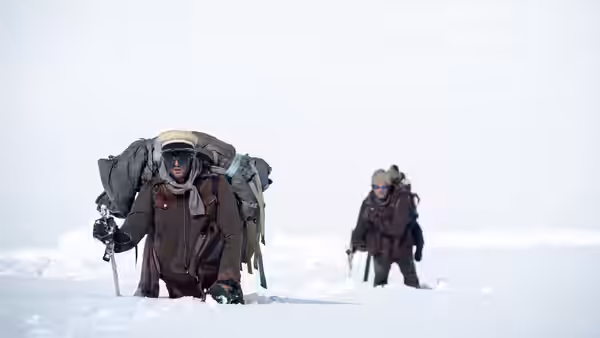 Two bundled survivors trudge through deep snow carrying large packs