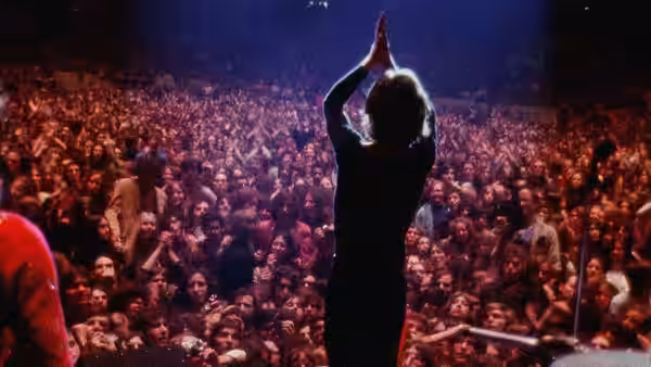 A performer stands with arms raised facing a huge crowd in an arena