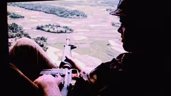 A soldier aims a machine gun from the open door of a helicopter above fields