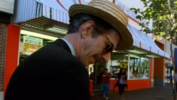 Robert Crumb in a straw hat walks along a city sidewalk