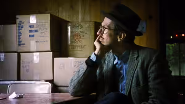 Robert Crumb in a fedora sits among stacked storage boxes in thought