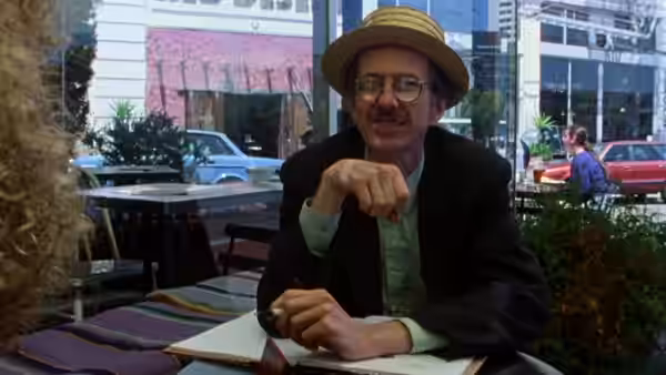Robert Crumb in a straw hat gestures while seated at a cafe table