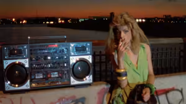 A woman in a green top sits beside a boombox on a waterfront at sunset