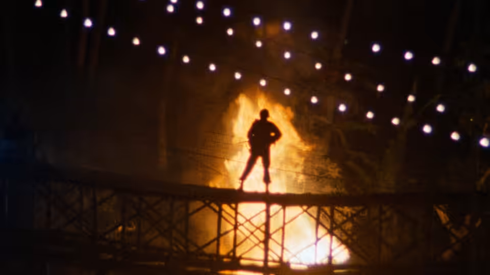 Silhouetted figure stands on metal bridge above bright flames and lights