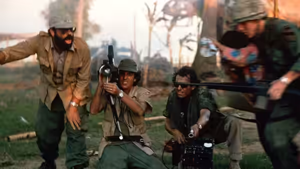 Bearded man directs while crew members hold cameras and rifles outdoors