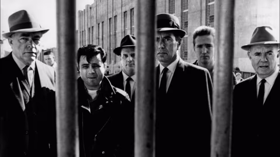 Several suited men stand behind vertical bars outside a large institutional building