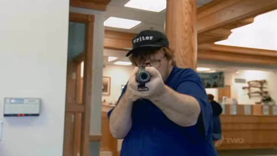 Michael Moore in a blue shirt and cap aiming a handgun with both hands indoors