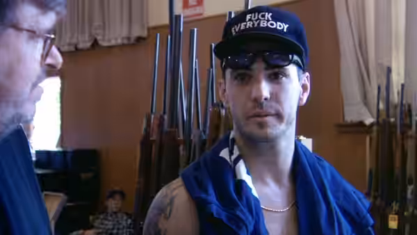 Man in a cap and sunglasses standing shirtless before a rack of rifles